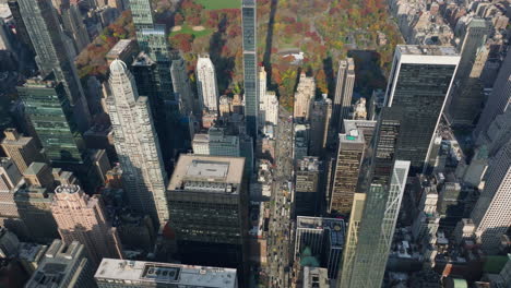 Forwards-fly-high-above-city.-Slowly-moving-cars-on-wide-avenue-between-downtown-skyscrapers.-Autumn-colour-trees-in-park.-Manhattan,-New-York-City,-USA