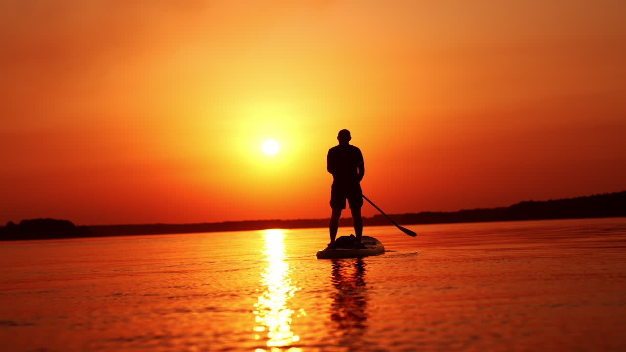 Man stands on the sup board facing the sunset. Man holds a paddle and row with it from each side.