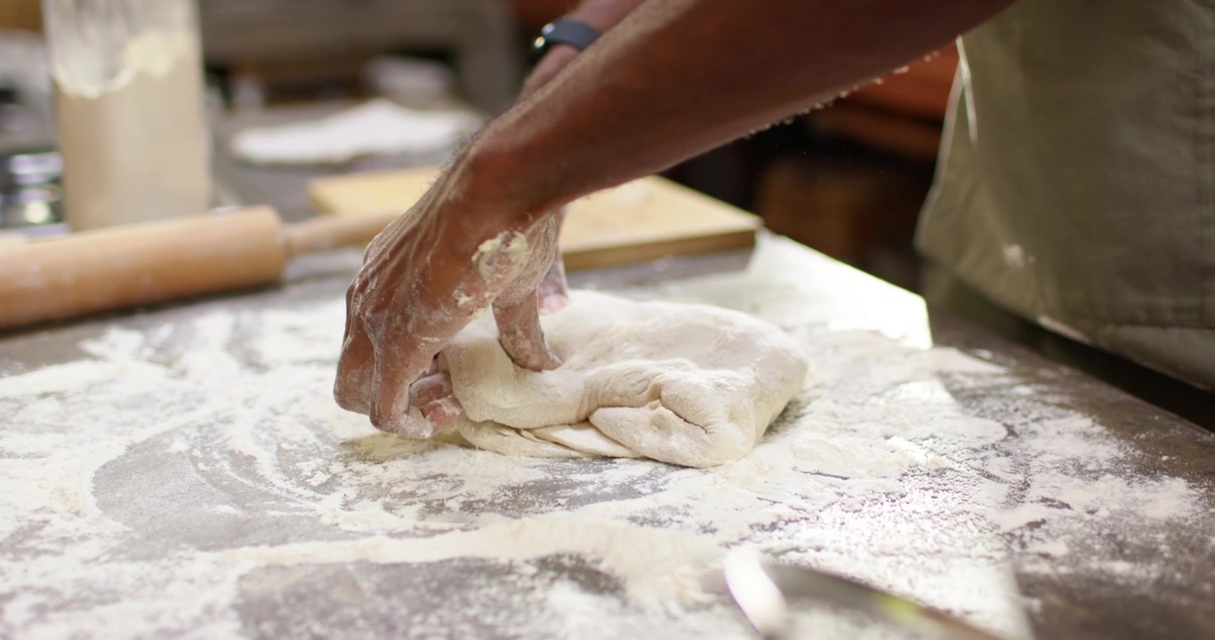 Male cook in apron from loose dough folding pressing at floured-counter near rolling pin for baking