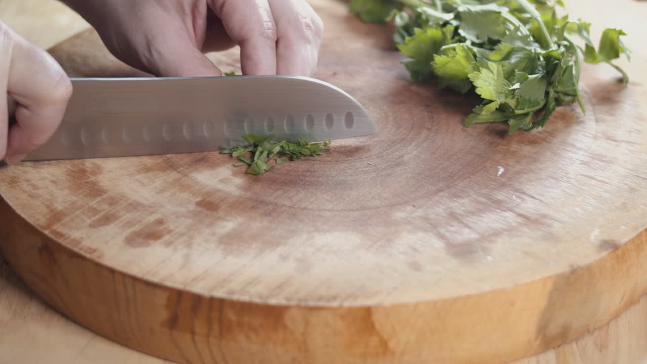 Slow Motion Slider Shot of a Chef's Hands Chopping Parsley With a Sharp Chefs Knife on a Thick Wooden Chopping Board Inside a Kitchen