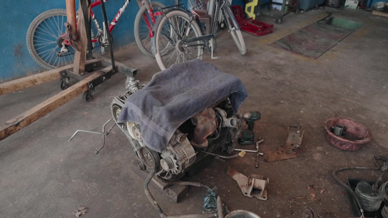 Removed car engine draped with cloth on a garage floor surrounded by tools and old bicycles