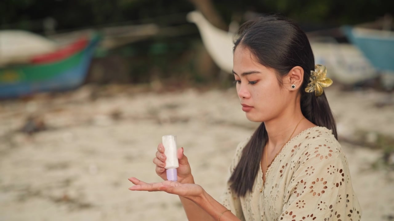 Asian woman applying sunscreen on beach, sitting on sand near colorful fishing boats, patterned dress and flower in hair, gentle breeze and golden sunlight, careful skincare ritual on hands and neck
