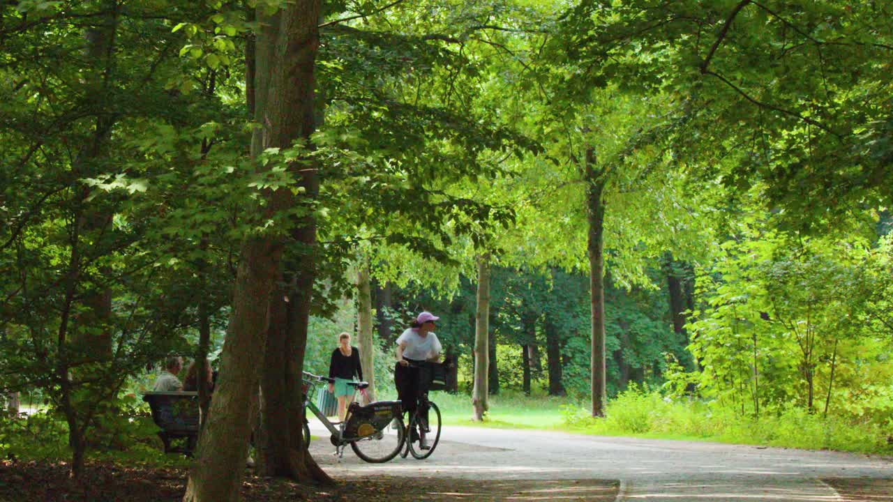 Two cyclists ride along a shaded, leafy park path in bright summer daylight, camera stationary