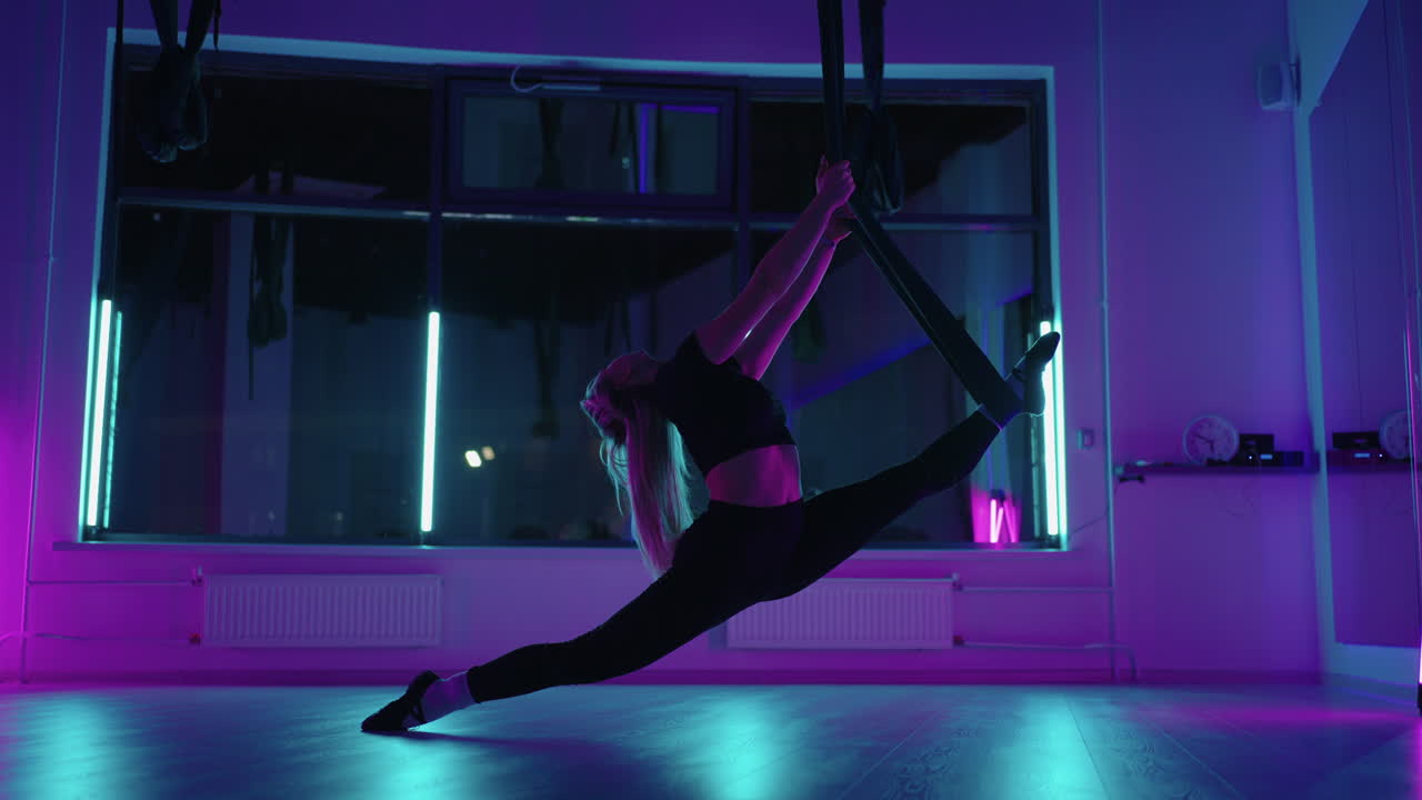 A woman does sports yoga in the air on a hammock in neon light performing graceful exercises and movements from stretching and dancing in slow motion.
