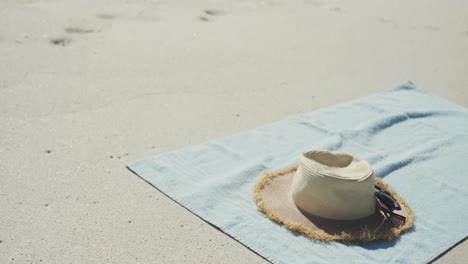 Close-up-of-straw-hat,-sunglasses-and-towel-on-beach,-in-slow-motion,-with-copy-space