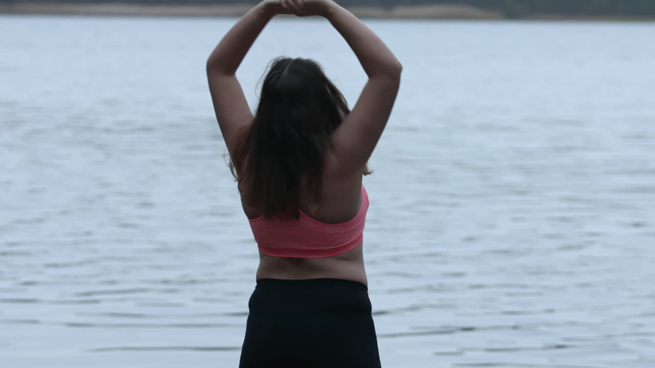 Happy overweight young woman on diet exercises, stretching next to a lake outdoors. Weight loss and active lifestyle concept