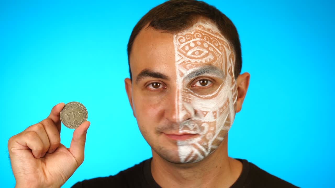얼굴이 칠해진 남자가 카메라를 바라보고 '예'라는 글이 새겨진 동전을 보여주고 있습니다.