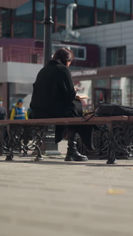 공공장소에 앉아서 책 페이지를 뒤집고 있는 여자의 뒷모습, 바람에 부드럽게 흔들리는 꽃, 바다를 가로질러 보이는 상점과 표지판을 가진 보행자