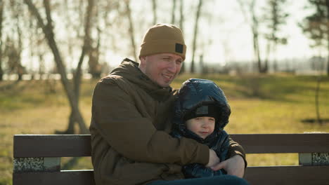 father and son sitting together on a park bench, with the son in a black jacket and the father in a brown jacket, surrounded by the blurred background of trees