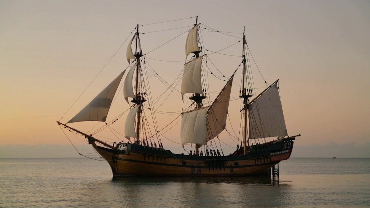 Veliero in legno a tre alberi che ondeggia sul mare costiero al tramonto, con le vele che si muovono per una leggera brezza.