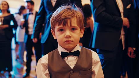elegant pageboy wearing formal vest and bow tie standing still in the foreground while blurred guests are dancing in the background at a wedding reception