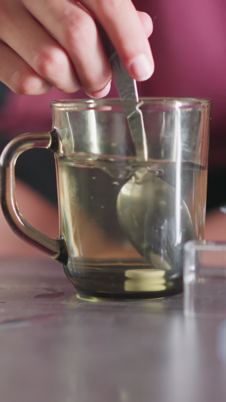Close up of young girl preparing medicine by pouring drug into warm water in glass cup with spoon nearby while seated on couch caring for sick brother during home treatment