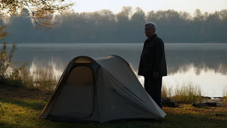 man standing next to a tent by a misty lake in the morning