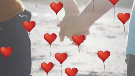 animation of red heart love balloons digital icons over couple holding hands on beach