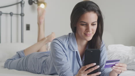 woman lying on bed with mobile phone shopping online using credit card wearing pyjamas