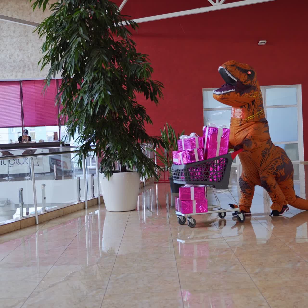 Man in the dinosaur costume is rolling the cart full of presents. Orange spotty dinosaur doing the shopping in the big mall