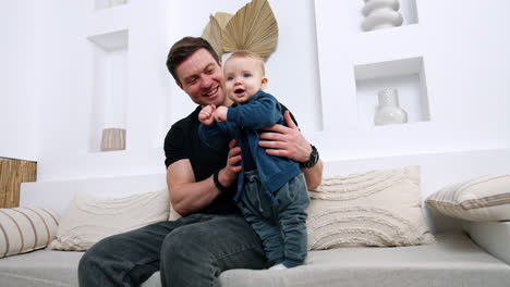 happy caucasian man sits on the sofa holding a baby boy. smiling cute baby stands supported by father. low angle view.