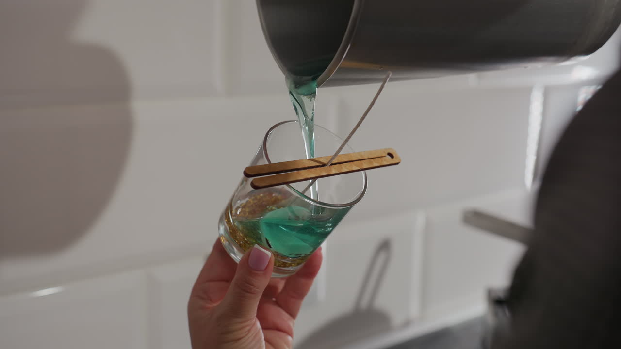 Craft person pours blue liquid into glass cup with two wooden sticks holding wick, preparing to make candle, close up of candle-making process in kitchen with modern appliance