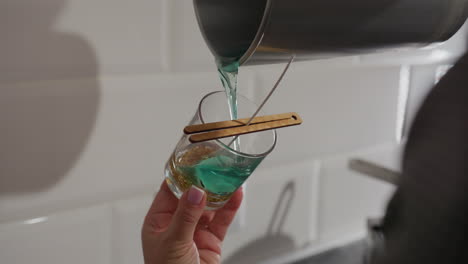 craft person pours blue liquid into glass cup with two wooden sticks holding wick, preparing to make candle, close up of candle-making process in kitchen with modern appliance