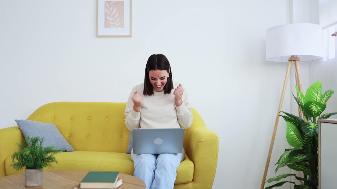 Mujer joven feliz celebrando el éxito mientras usa el portátil en el sofá