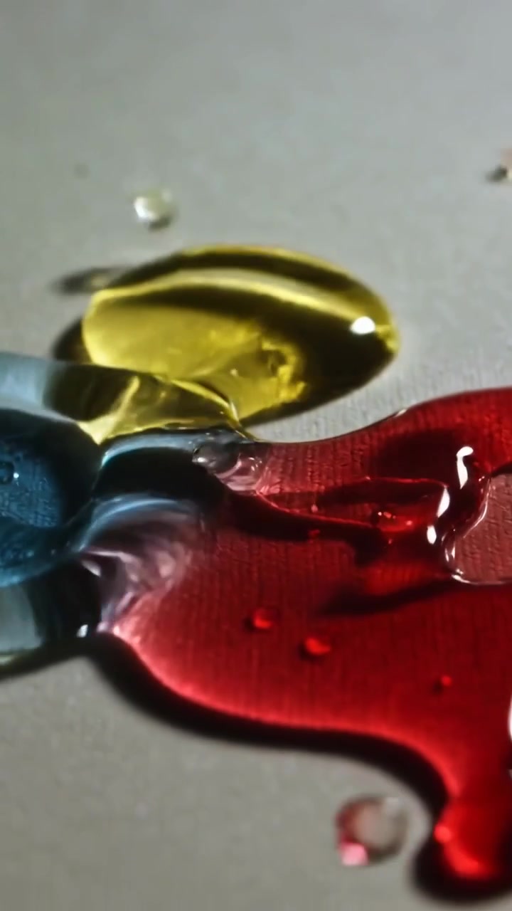 Vertikales Video: Ein aufprallender roter Tropfen erzeugt blaue, gelbe Pfützen, die sich auf einer grauen Matte ausbreiten, mit kleinen Perlen.