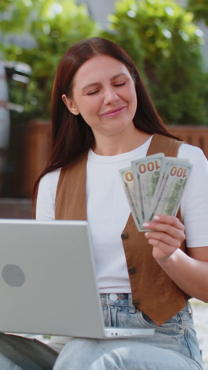 Mujer rica feliz usando portátil ganador victoria sosteniendo dinero en efectivo en billetes de dólar en la calle de la ciudad
