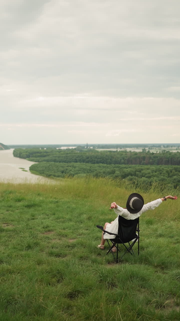 A woman in a black hat sitting comfortably on a chair, stretching her arms while an artist paints her in a lush grass field beside a tranquil lake