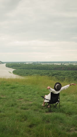 A woman in a black hat sitting comfortably on a chair, stretching her arms while an artist paints her in a lush grass field beside a tranquil lake