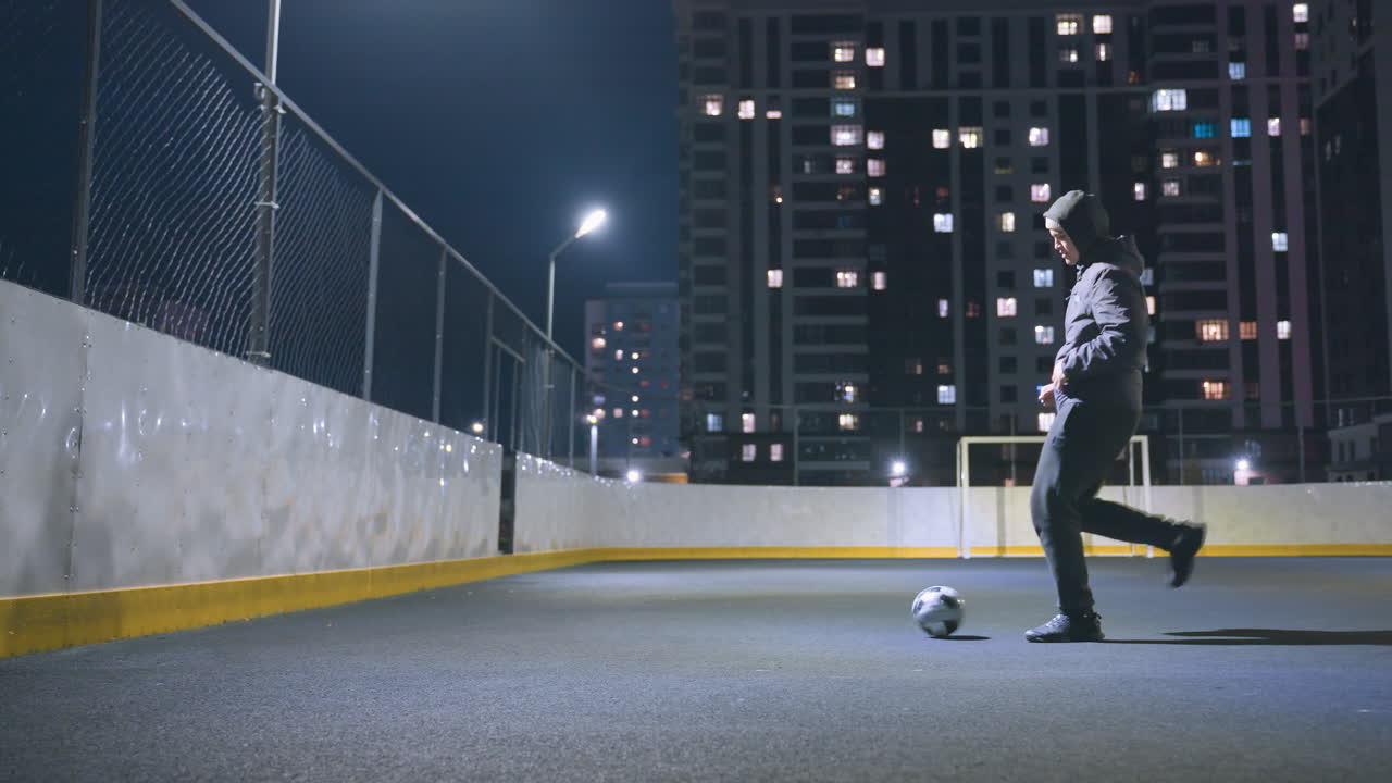 vista lateral de un hombre con capucha pateando una pelota de fútbol bajo la iluminación brillante del estadio por la noche, cerca del poste de la portería y reflejándose en la pared. paisaje urbano con edificios de oficinas iluminados