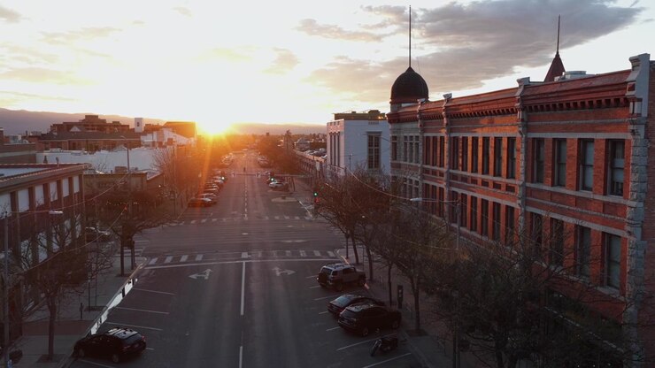 Via principale del centro storico di Missoula, Montana, al tramonto dorato, con classici edifici in mattoni, auto parcheggiate, alberi spogli che costeggiano una strada tranquilla e un cielo drammatico e luminoso con nuvole. Ripresa aerea in avvicinamento.