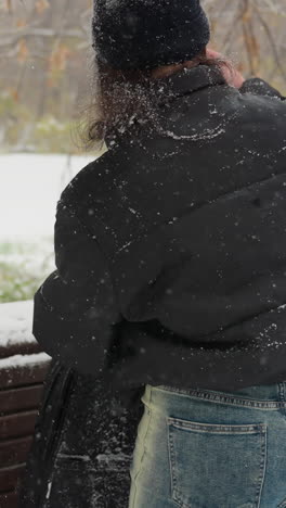 vista trasera de alguien abriendo un paquete de guitarras colocado en un banco de parque cubierto de nieve, con la nieve cayendo suavemente, una rama verde en foco y un paisaje de invierno sereno en el fondo