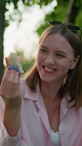 close-up of woman smiling while holding bread in hand and enjoying outdoor meal, seated in lush green park, with tattooed hand holding bread partially visible in background