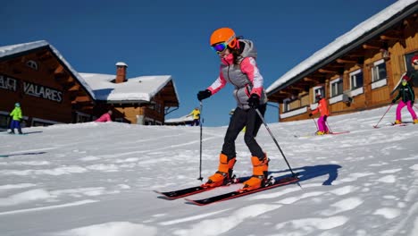 esquiador de invierno con casco naranja brillante y gafas de protección deslizándose suavemente por una ladera de montaña cubierta de nieve cerca de un chalet de madera, encarnando la libertad recreativa alpina y la energía de los deportes de invierno