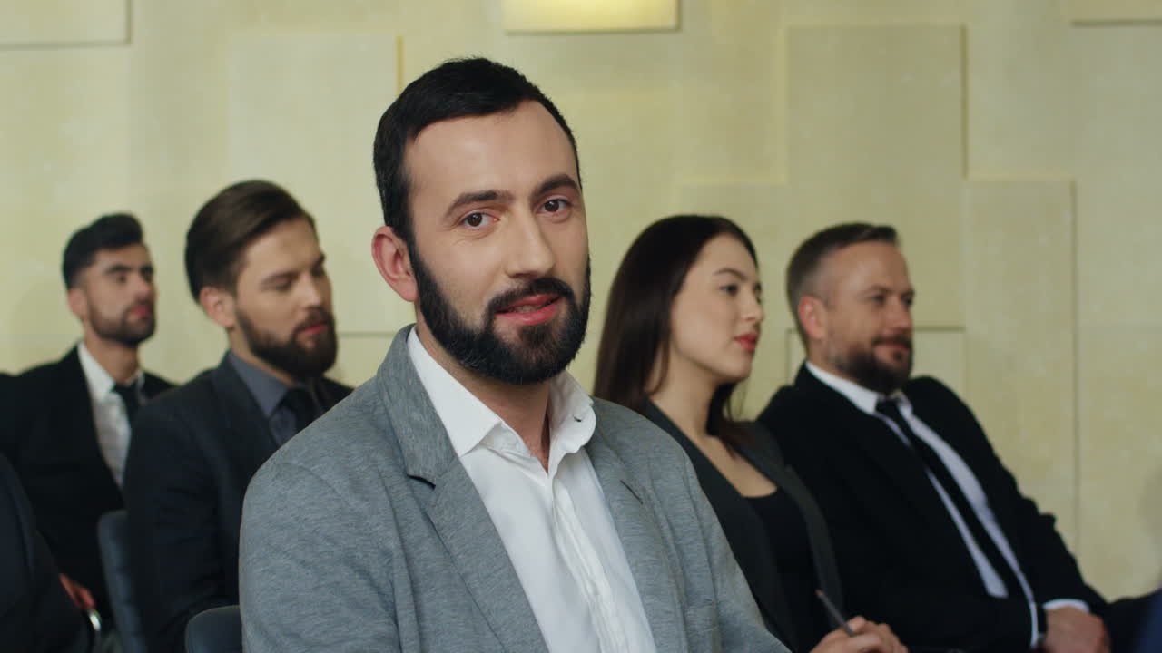 hombre de negocios caucásico sentado en una silla entre personas en una sala de conferencias, luego gira la cara hacia la cámara y sonríe