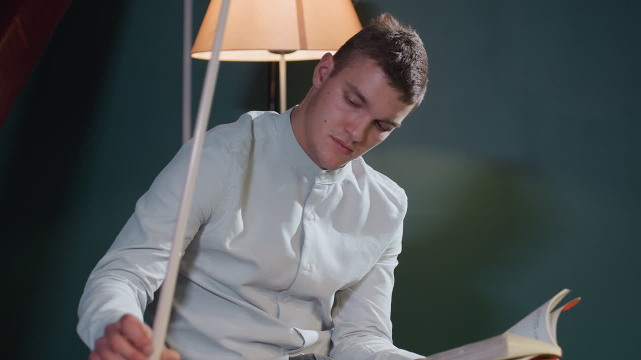 Close-up of guy in white shirt sitting and reading book while holding cue stick with focused expression. Warm lighting enhances intellectual and strategic atmosphere in modern indoor setting