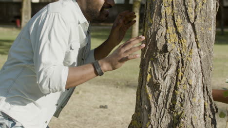 black man and playful boy hiding around tree, playing in park.