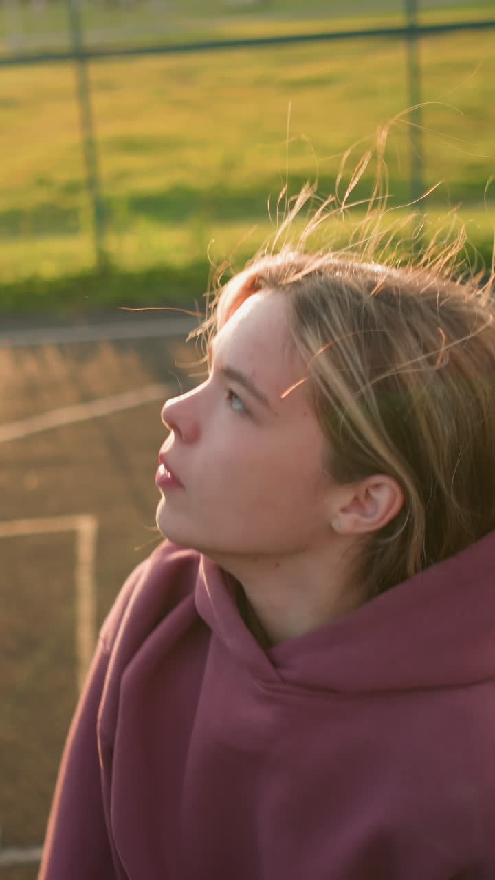 joven con capucha marrón rebotando voleibol, preparándose para servirlo en un campo abierto con marcas de la cancha en el suelo, el sol proyectando un brillo cálido, centrado en su próxima jugada