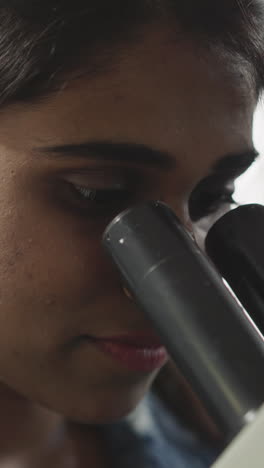 focused indian woman looks through microscope on blurred background. young scientist does analysis of test sample in medical laboratory closeup