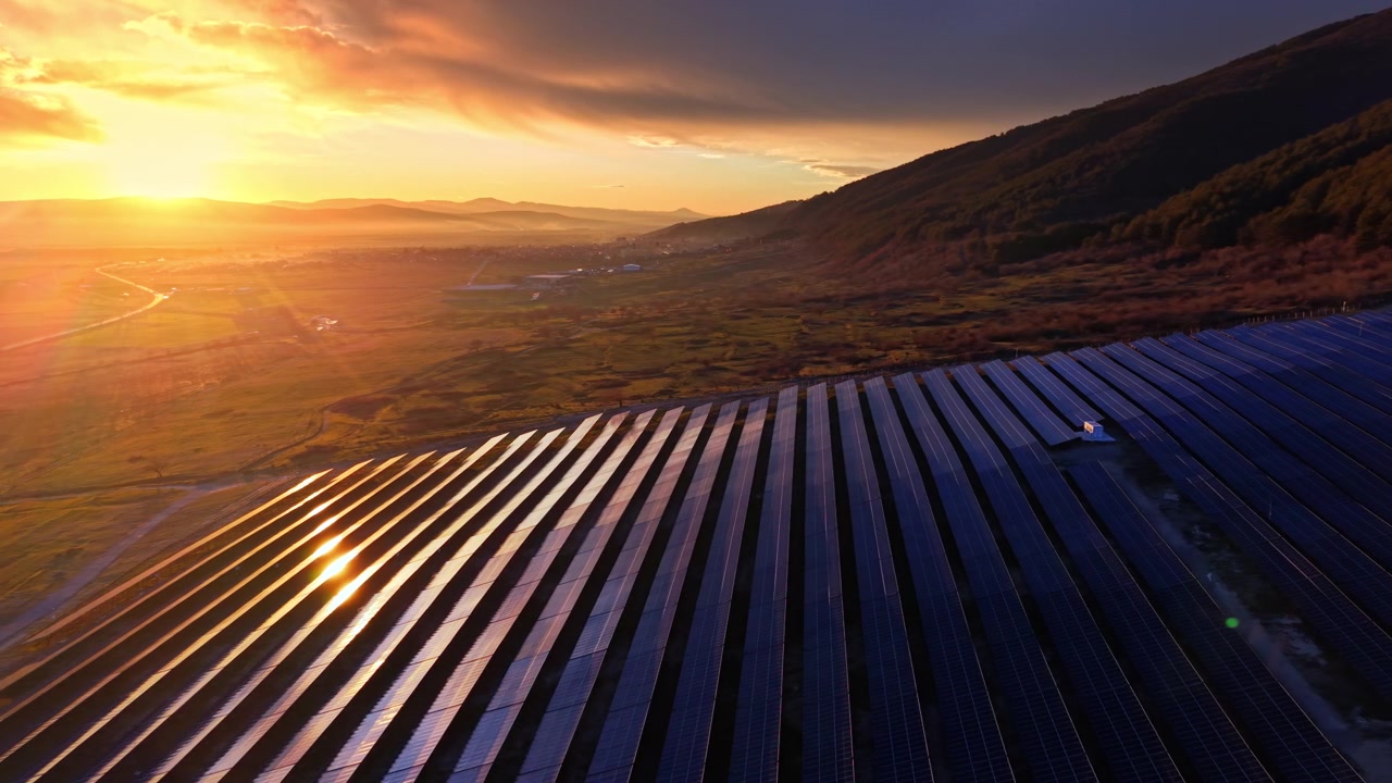 Solopgangsbillede af rækker af solpaneler over en dal. Luftfoto viser solglimt, der reflekteres fra panelerne, med en tåget landsby synlig nedenfor. Stort solcelleanlæg