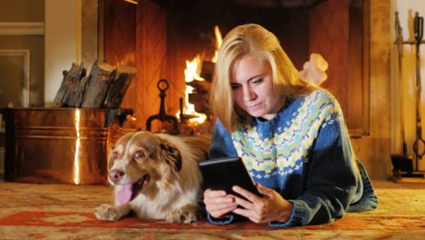 blonde young woman reading e-book reader against the background of a burning fireplace. next to it is a large dog