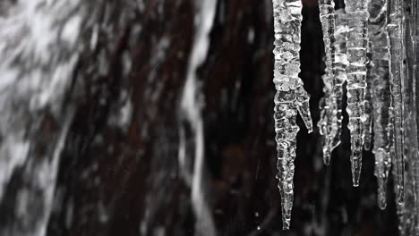 Wasser-Plätschert-An-Einem-Wasserfall-Mit-Schwarzen-Felsen,-Während-Im-Hintergrund-Schneeflocken-Herabwirbeln-Und-Lange-Eiszapfen-Herabhängen