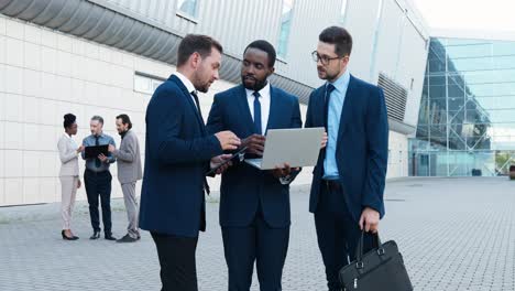 three caucasian and african american businesmen in stylish clothes reading something on the laptop and talking in the street