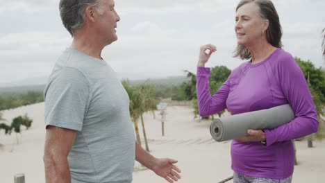 Portrait-of-happy-senior-caucasian-couple-holding-yoga-mat-and-embracing-on-beach,-in-slow-motion