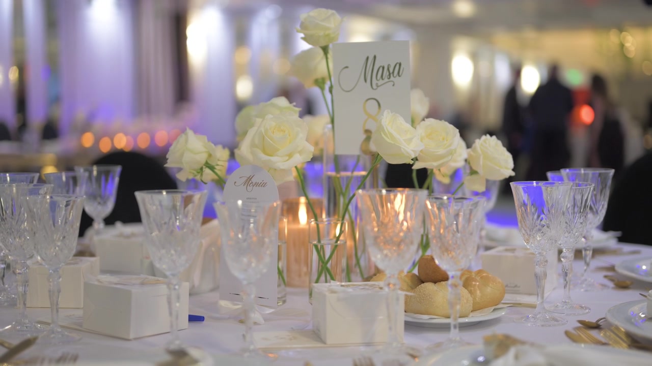 Allestimento elegante di un tavolo da banchetto con rose bianche, bicchieri di cristallo, candele e menu decorativi in un ambiente interno con luce soffusa.