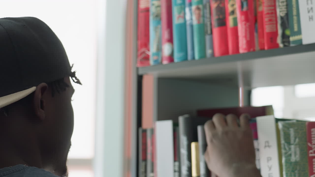 Close up rear view of young man wearing black cap browsing bookshelf in library, hand reaching out to select book among colorful spines, blurred background