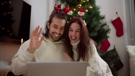 Happy-guy-and-girl-in-New-Year's-hoops-and-white-sweaters-sit-in-front-of-a-gray-laptop-and-congratulate-their-friends-and-family-Happy-New-Year-near-the-New-Year-tree-in-a-cozy-room-in-a-winter-evening