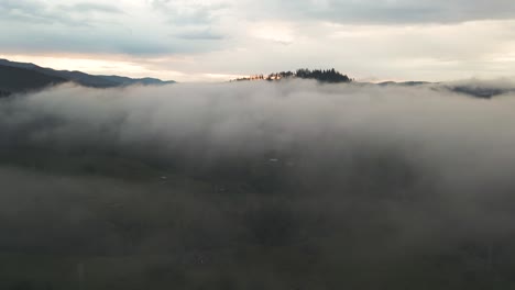 Flying-over-landscape-covered-by-fog-towards-a-forested-peak,-sunrise-barely-visible-above-the-horizon