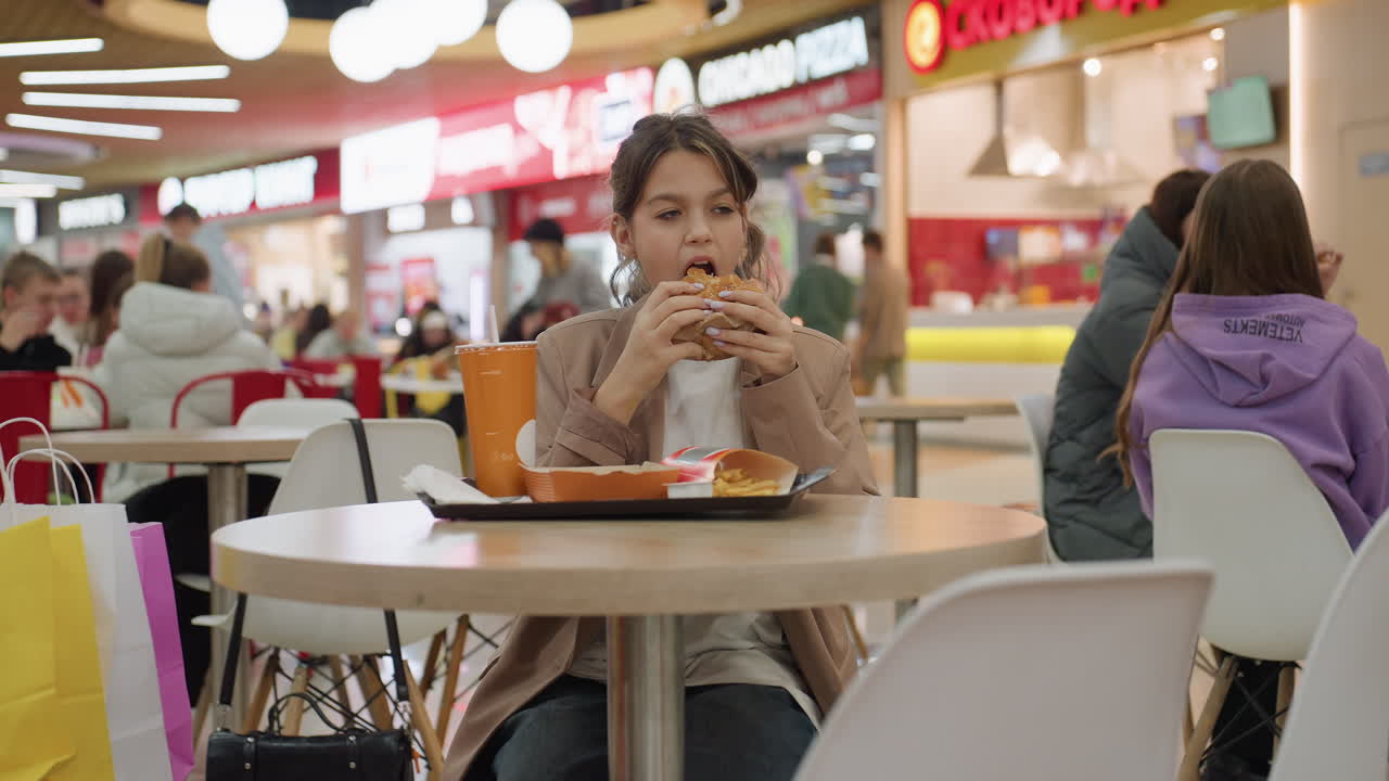 Joven comiendo en medio de compradores ajetreados, estudiante saboreando una hamburguesa en un concurrido centro comercial, estudiante relajado con comida rápida rodeado de compradores bulliciosos y ambiente urbano.