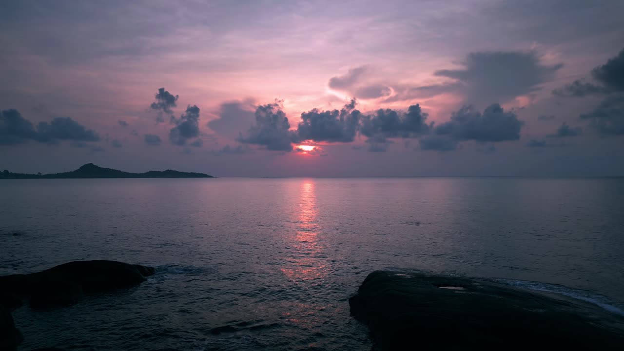 A serene seascape captured during sunset, featuring a sky filled with soft clouds glowing in hues of pink, purple, and gold. The partially hidden sun casts a warm, ambient light across the calm ocean