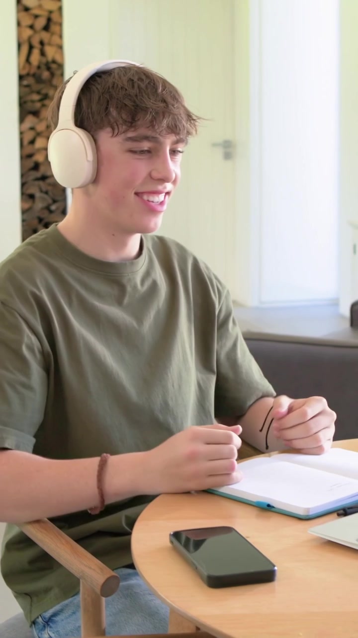 Vertical video: Hearing student with headphones at home taking notes on laptop, notebook for study